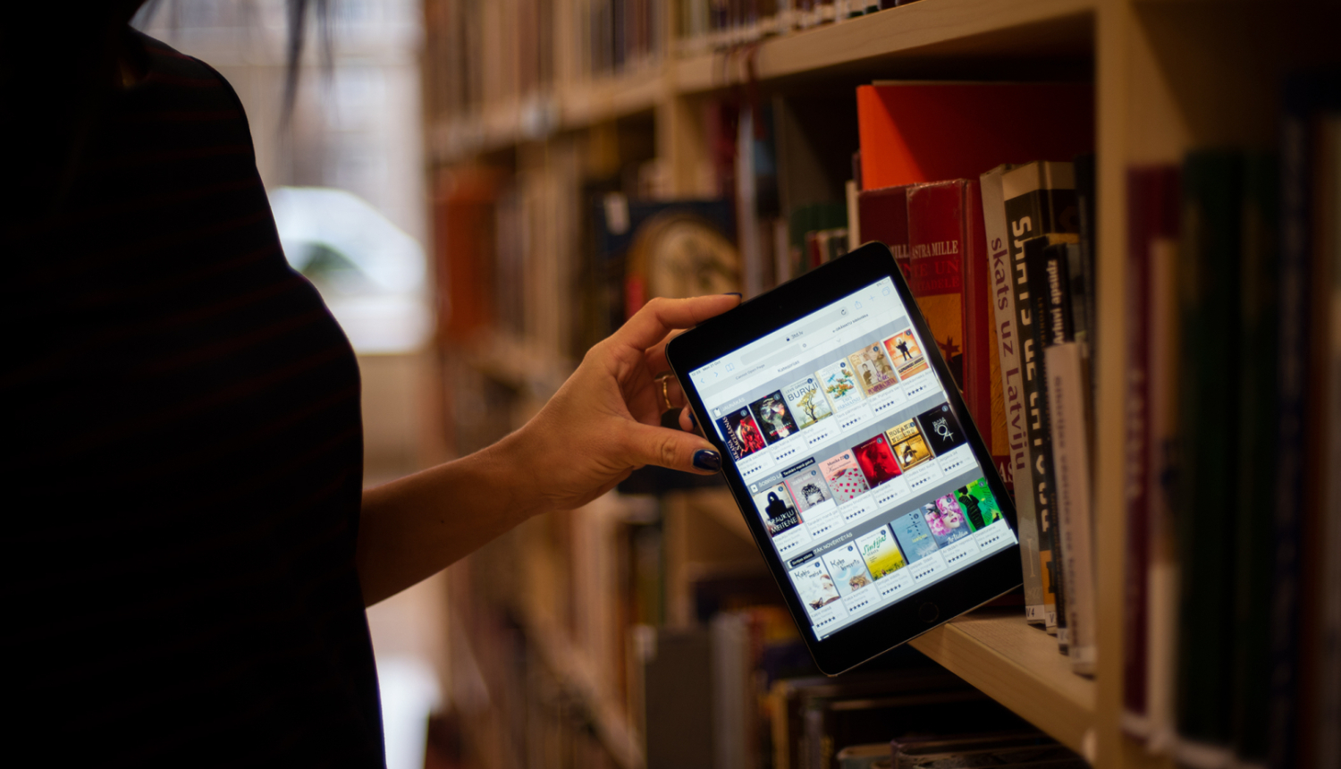 Sieviete ievieto bibliotēkas grāmatplauktā planšeti ar atvērtu 3td.lv skatu